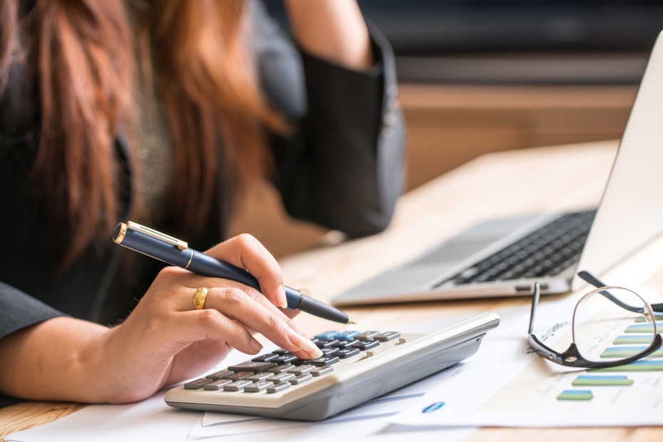 A woman sits at a desk in front of an open laptop. In her right hand, she holds a pen and punches numbers on a calculator.